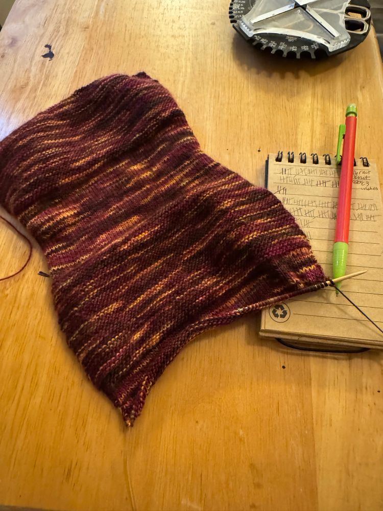 A piece of knit fabric on a table next to a notebook and pencil with hash marks on the paper and a knitting tool kit in the background.
