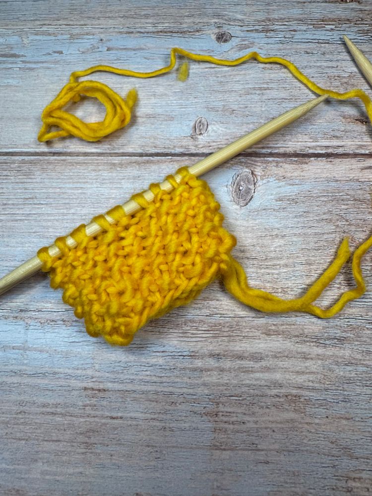A swatch of seed stitch worked in the round in bulky yellow yarn is shown on a wooden double pointed knitting needle.