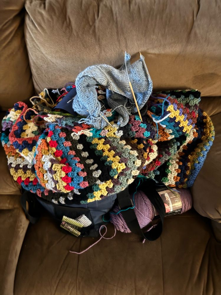 A beige couch seat covered with a bag full of yarn, a mostly done crocheted blanket, a piece of blue knitting and a piece of blue cross stitch.