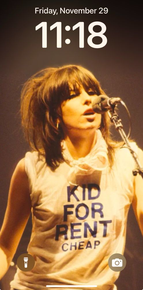 Screenshot of my lock screen. Young Chrissie Hynde of the band The Pretenders. She has mid length brown hair teased up in the back, and bangs cut low almost covering her eyes with heavy black eyeliner. She is leaning into a microphone on a mic stand, mid lyric. She wears a white t-shirt with sleeves rolled up. The shirt reads KidFor Rent in large black letters. Under those words in smaller black letters it says Cheap. She is a rock goddess 