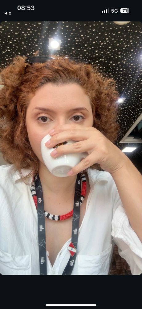 Mulher branca e ruiva de cabelo cacheado/ondulado. Ela toma um cafezinho olhando pra câmera.
Usa cordão de crachá com pin do Pará, além de um colar estilo cobra coral