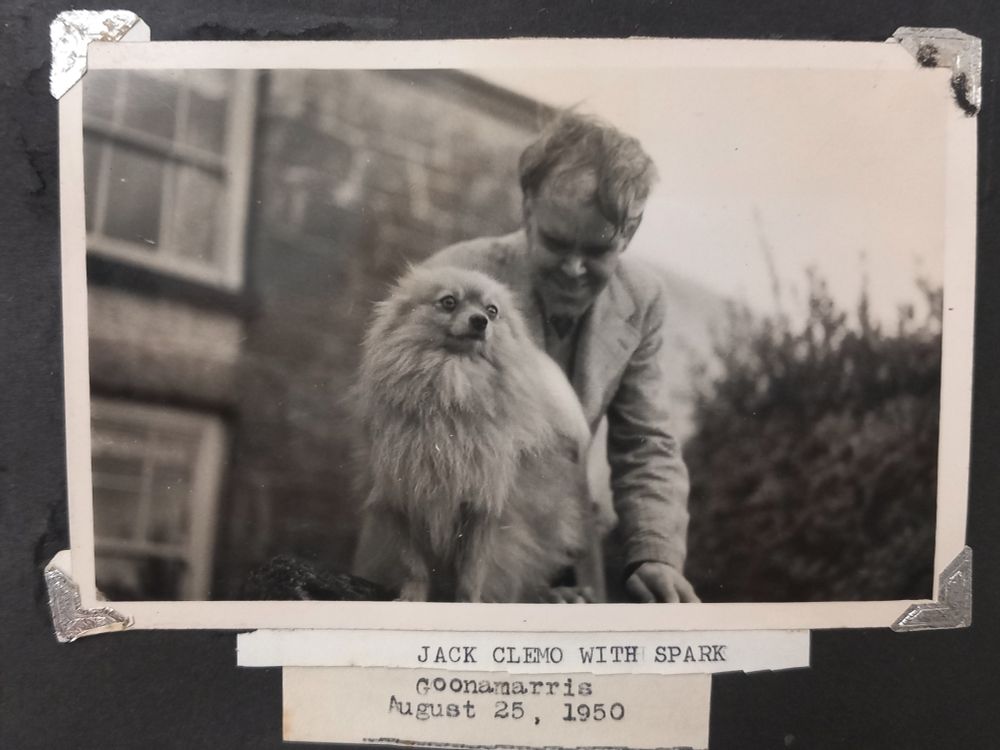 Cropped image from a black photograph album page showing a black and white photograph of Jack Clemo leaning over a light-coloured Pomeranian in the foreground. The dog looks majestic, but his eyes look slightly alarmed! In the background is the cottage Goonamarris. The typed caption underneath reads: 'JACK CLEMO WITH SPARK Goonamarris August 25, 1950'.