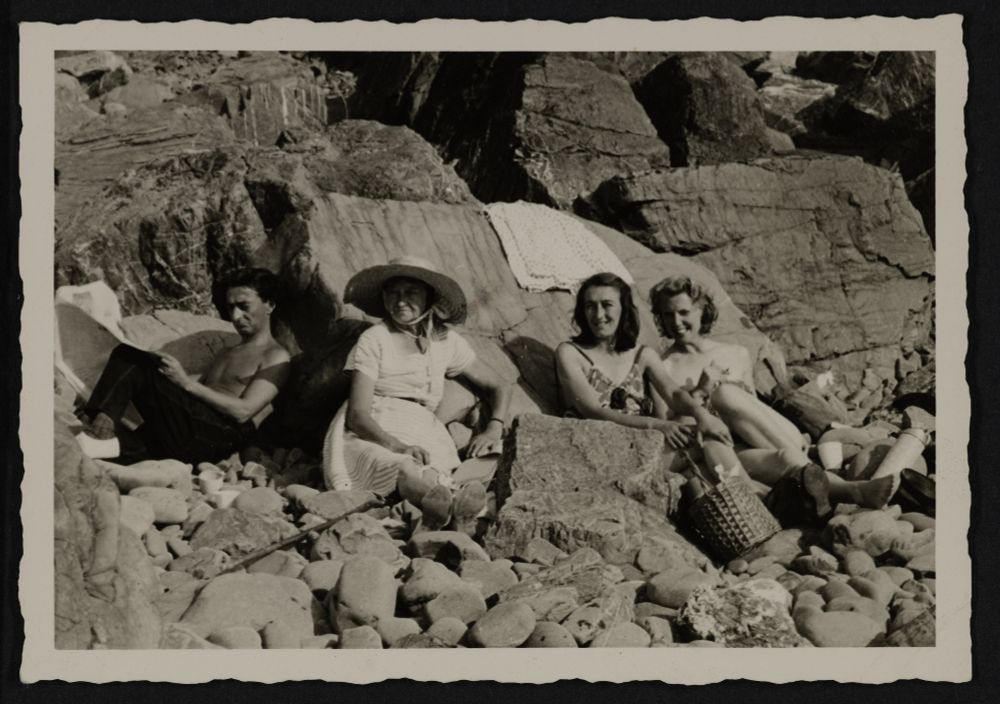 Black and white photograph taken on a pebbly beach with large rocks in the background. On the left is Ronald Duncan, who is frowning and seated facing sideways. Next to him are seated three women (his mother, Mole, his sister, Bunny, and his wife, Rose Marie), who are smiling and looking at the camera. A picnic basket can be seen in front of them. 