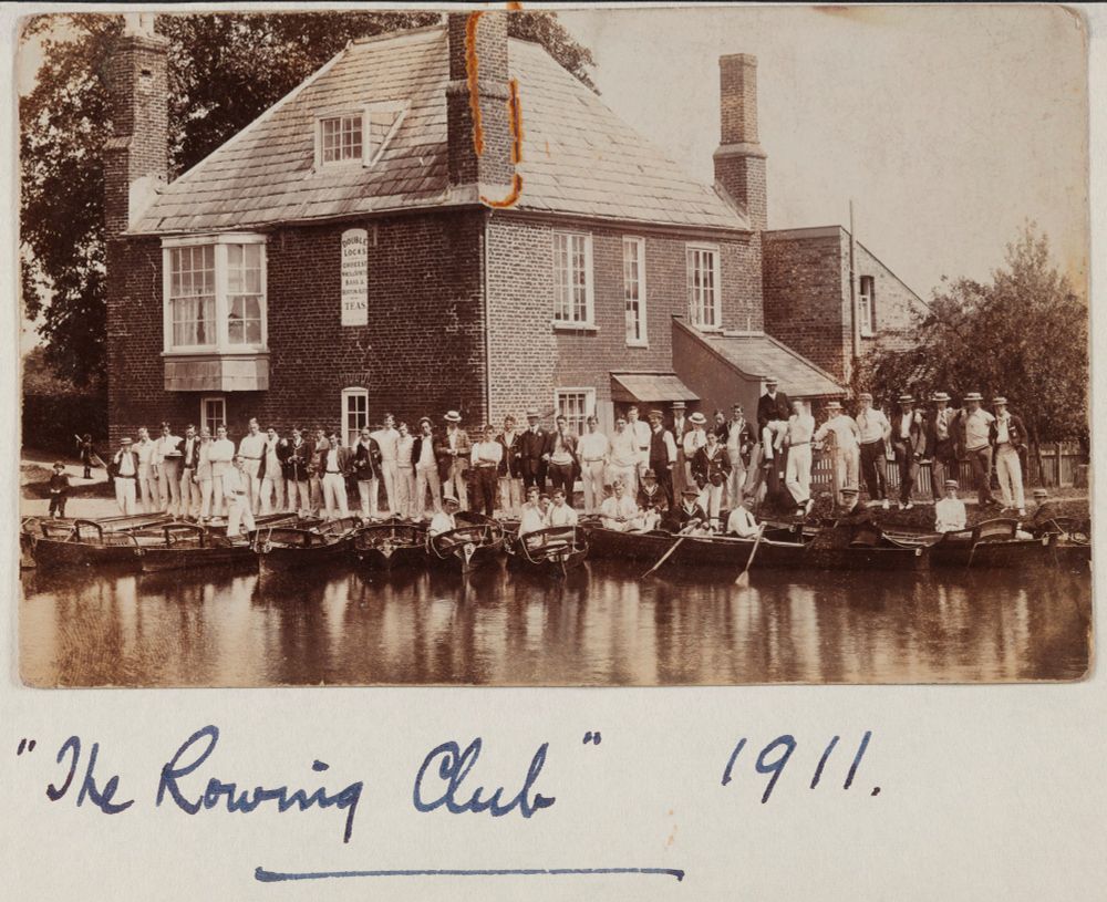 Sepia-toned photograph of a long row of students standing outside the Double Locks pub. Several rowing boats are moored in front of them, and some students are sitting in the boats. Two young children are also visible on the left hand side. The caption underneath reads '"The Rowing Club" 1911.'. A rusty paperclip has left a mark at the top of the photograph.