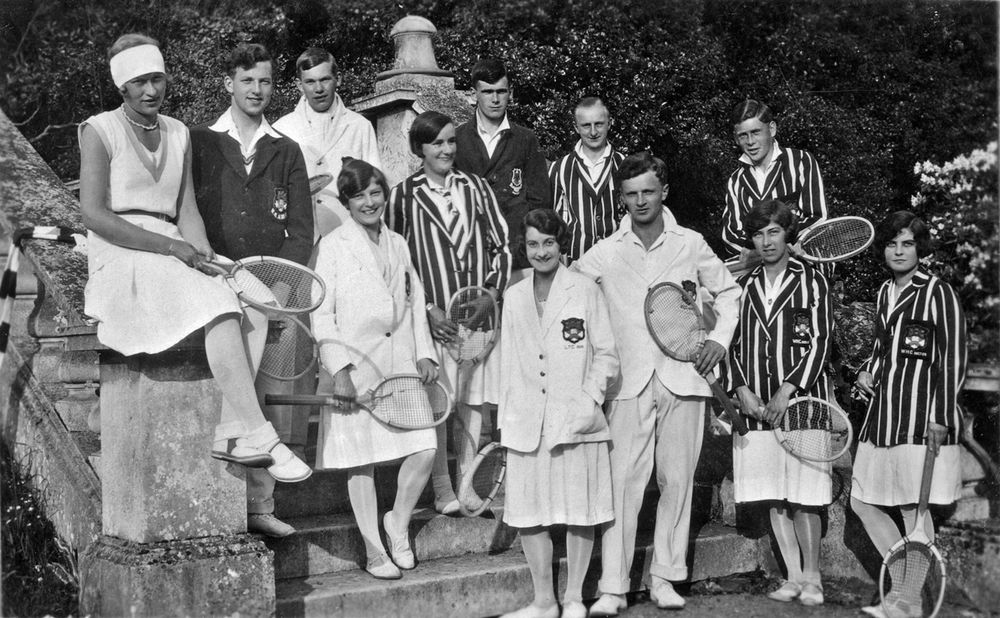 Black and white photograph showing twelve students - six women and six men - on and around stone steps within the grounds of Reed Hall. Five students are wearing white blazers; two are wearing dark blazers, and five are wearing striped blazers. All are holding tennis rackets.