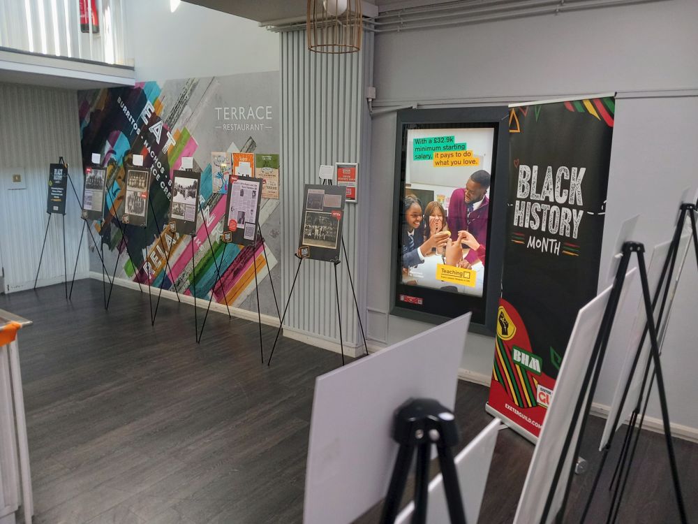 Pop up exhibition of boards on easels. Each easel has an information card and the boards feature archival photographs and newspaper clippings. Each board represents a different time period at the University, beginning with the 1940s. In the middle of the exhibition is a big banner for Black History Month.