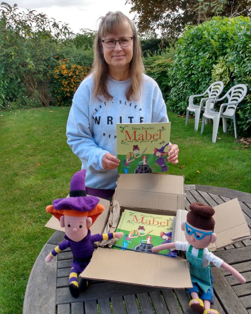 Photo of Author Dawn Treacher holding a copy of Mabel and the Flower Spell in her garden with a box of books in front of her and plush toys of the two lead characters.