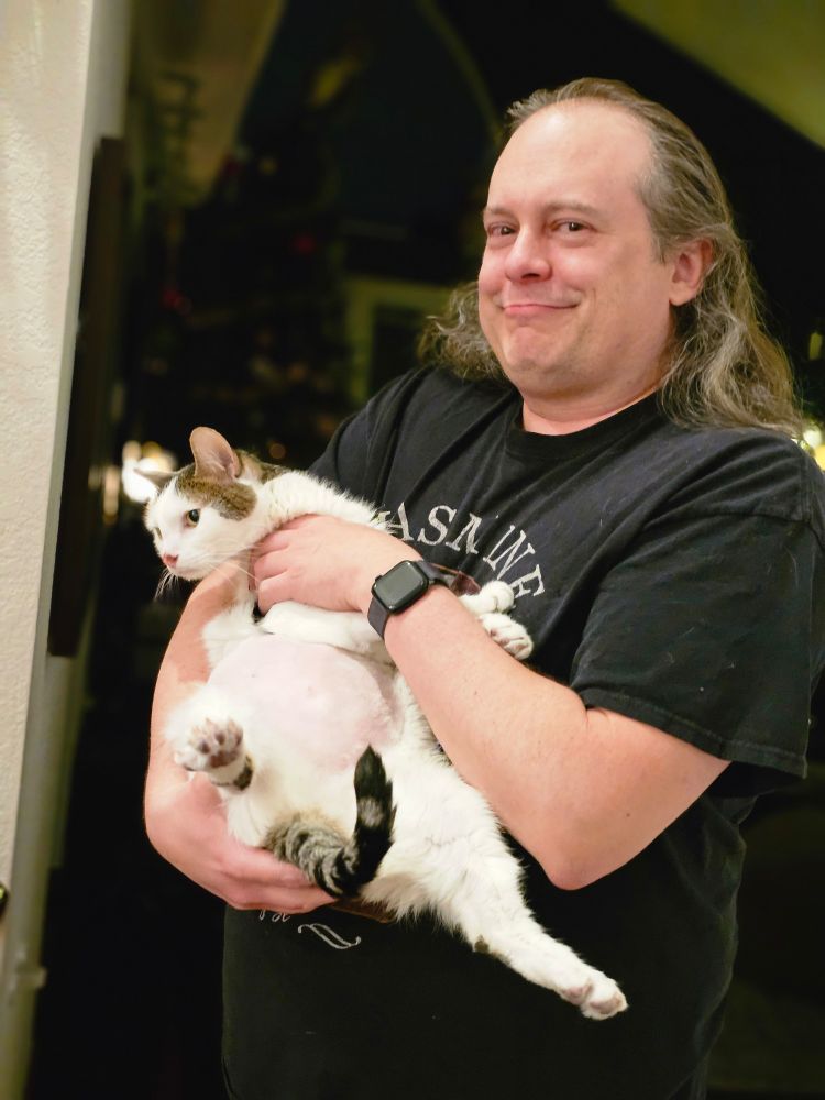 A adult man smiles lovingly while holding a white cat whose shaved belly is visible. The cat's legs are sticking out with toes spread as the cat wriggles to get free of the human's grasp.