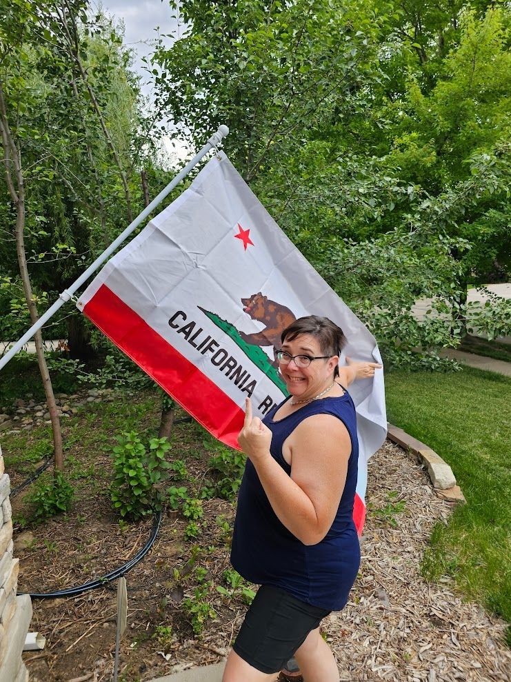 Photograph of original poster, @stsunflower.bsky.social‬, giving the middle finger to the camera while posing holding a California state flag that's flying outside a residence on a summer day