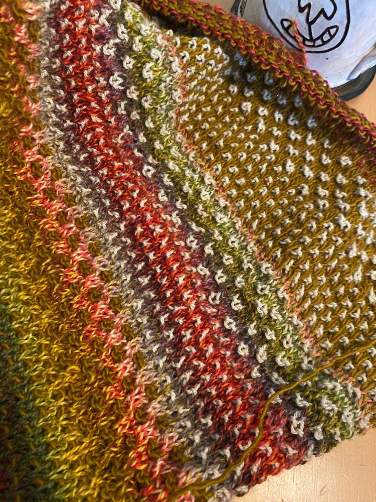 A hand knit piece of multi-colored fabric lies across a wooden table. Yarn colors include greens, orange, reds, pinks, and white. A mug with an image of a smiling dog face is in the background of the picture. 
