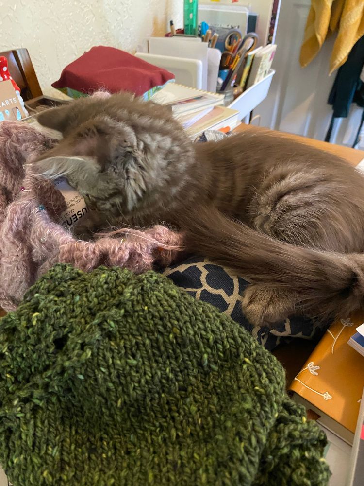 A brownish gray kitten lays on a messy table on top of a pile of knitting with her head and front legs stuck deep into one of the knitted projects.