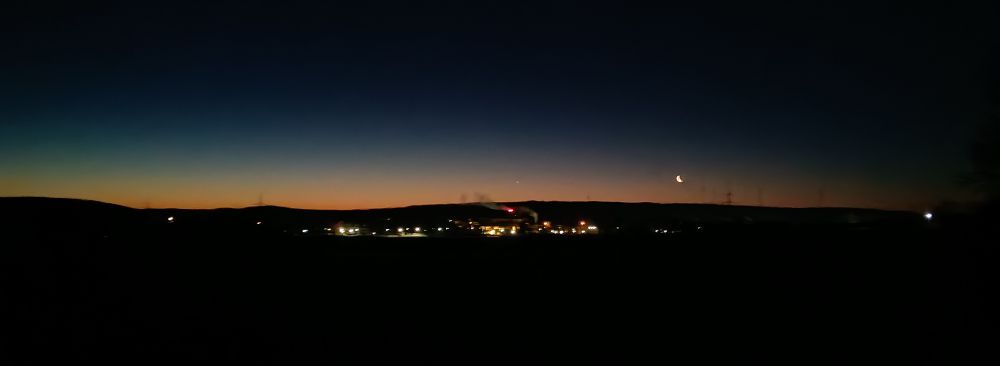 Hinter im Dunkel liegenden Wiesen sueht msn ein paar Lichter des Städtchens. Direkt über dem Horizont verfärbt sich der Nachthimmel leicht orange und die schmale Mondsichel leucht.
