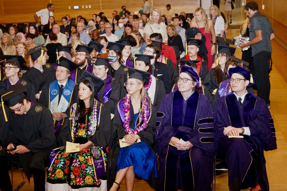 Students in cap and gown sitting at Graduation with supporters in the background.