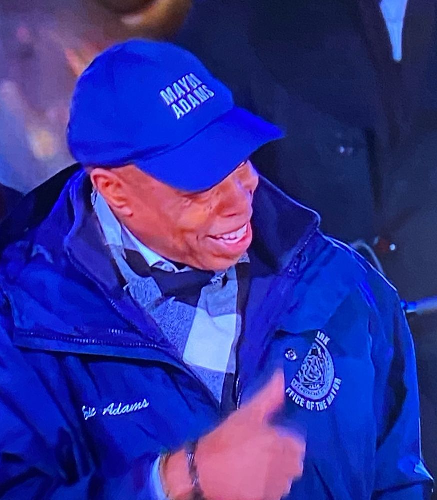Eric Adams on TV wearing a big stupid hat that says ERIC ADAMS on it, giving a thumbs up