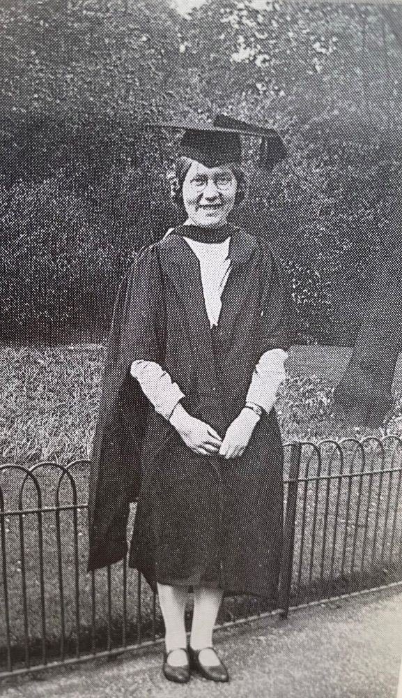 a b&w photo of Elsie Widdowson as a young woman on one of her graduation days. She wears a gown and mortar board with a while blouse. She is smiling happily and stands on a park with trees and shrubs behind