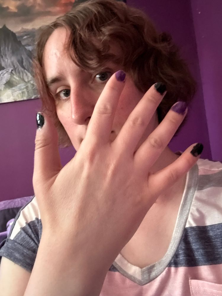 Ruby holds her hand in front of her face, her nails painted in a sequence of alternating black and purple. she is wearing a bright pink striped shirt 