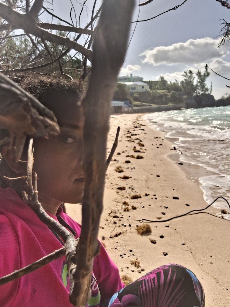 Woman on the beach, colourful workout clothes. Her face is obscured by branches in the foreground. 