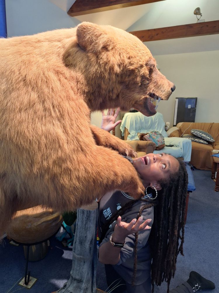 A black woman with long locs pretending to be attacked by a taxidermied brown bear. Her arms are up in faux defense and she's fake yelling. 