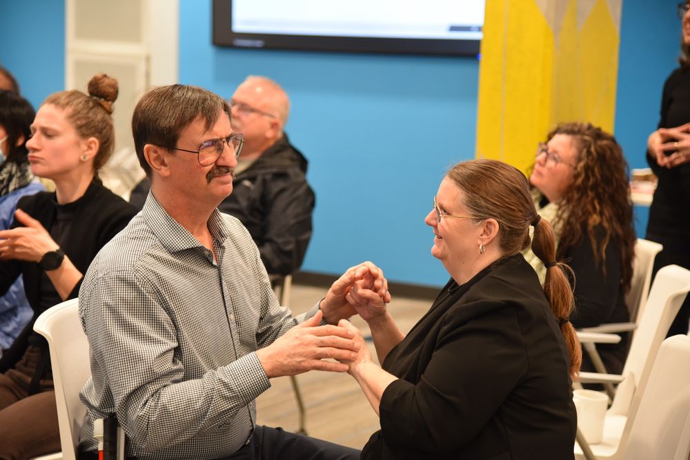A man and woman sit facing each other, smiling and using tactile sign language to communicate. Other participants in the background are also engaged in various forms of communication during a community gathering.