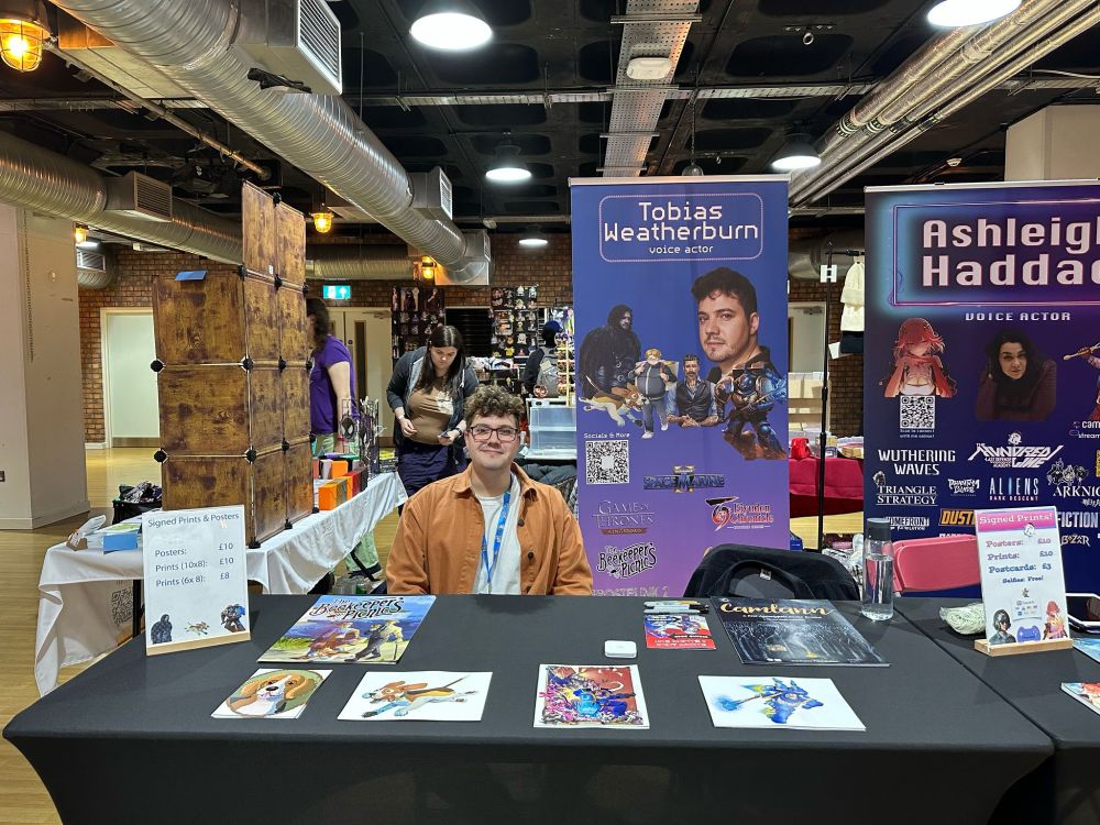 Tobias Weatherburn, a voice actor with curly hair and glasses weating an orange shirt over a white top, sits behind a signing table in a convention hall. The table has posters and prints from Space Marine II, The Beekeeper's Picnic, and Camlann on it. Behind the table to the right of Tobias is a purple banner with Tobias's name and face on it, as well as a list of credits and pictures of some of the characters he has voiced