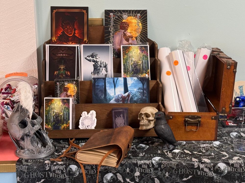 A 4 tiered display shelf with an art print of Karlach (Baldur’s Gate 3) and Kiriona Gaia from The Locked Tomb at the top. In 2nd shelf is a print of the Lotus and The Man in the Wall (Warframe), a stone statue cracked with gold liquid coming from it, and a statue of a dnd character with skeleton wings as they’re surrounded by flora. The 3rd shelf is a smaller print of Kiriona Gaia and another of a pink haired woman crawling from the ground with a blue moon behind her. On the bottom shelf is a smaller version of the dnd character print and a sticker of a skull with a sword and flowers going through it, as well as a business card. 