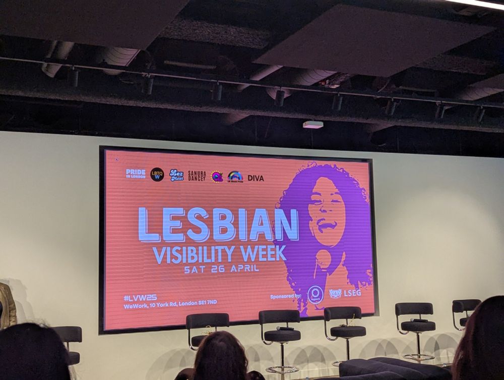A brightly coloured screen which reads Lesbian Visibility Week with an image of a smiling person with curly hair. Behind several tall chairs in an auditorium 