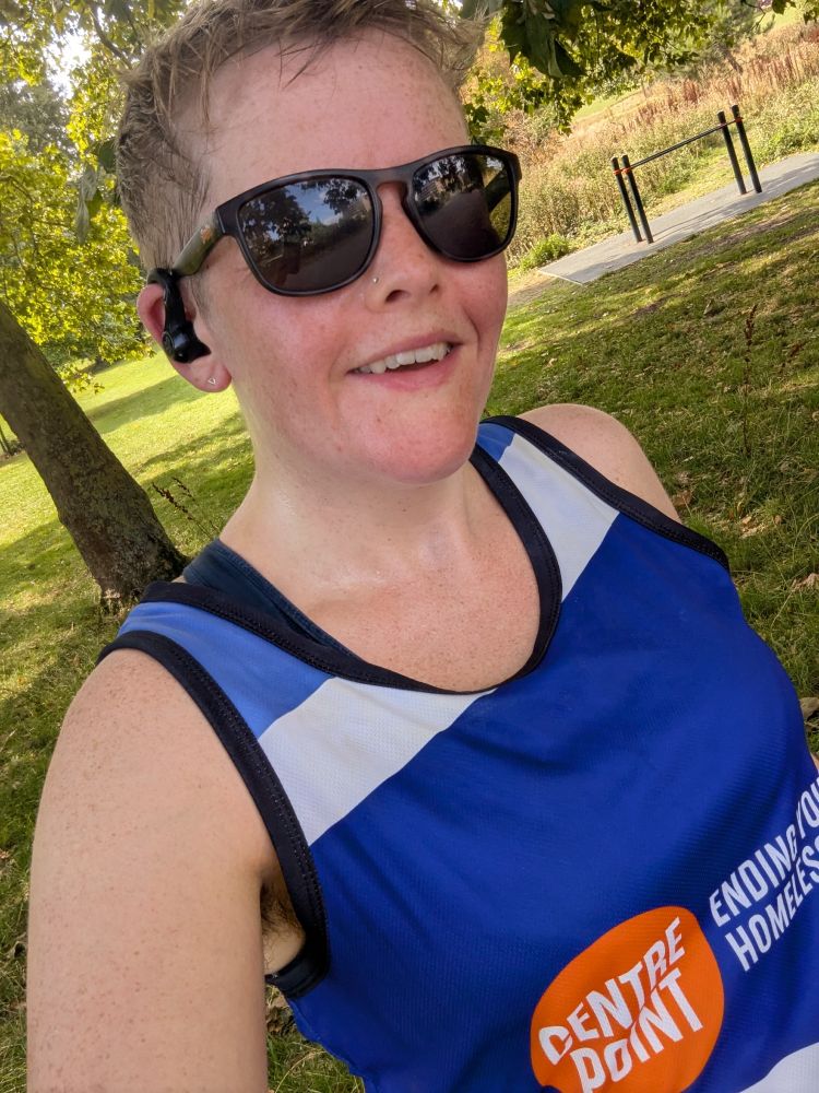 I stand in a sunny park, in the shade of a tree. Wearing sunglasses and my Centrepoint running training top and sunglasses.
