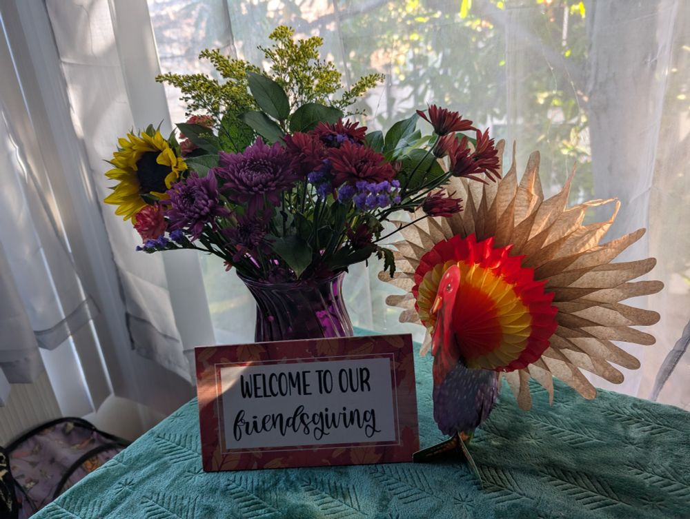 A bouquet of flowers with a pop open paper turkey decoration and a sign: "welcome to our friendsgiving"