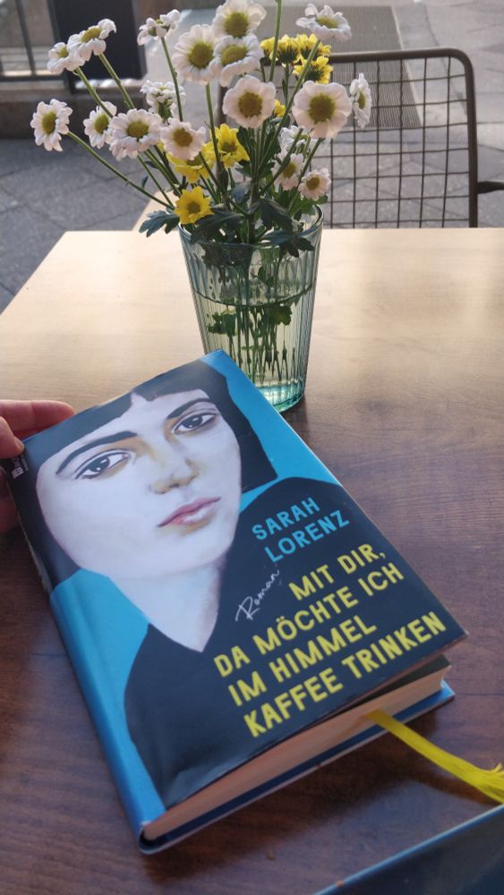 Der Roman "Mit dir, da möchte ich in Himmel Kaffee trinken" von Sahra Lorenz liegt auf einem Tisch. Auf dem Tisch steht Glas mit weißen und gelben Blumen.  Das Buchcover zeigt eine weiße Frau vor blauem Hintergrund, der Buchtitel ist in gelb geschrieben. Eine Hand hält das Buchcover leicht geöffnet. Der Tisch steht auf einer Straße.