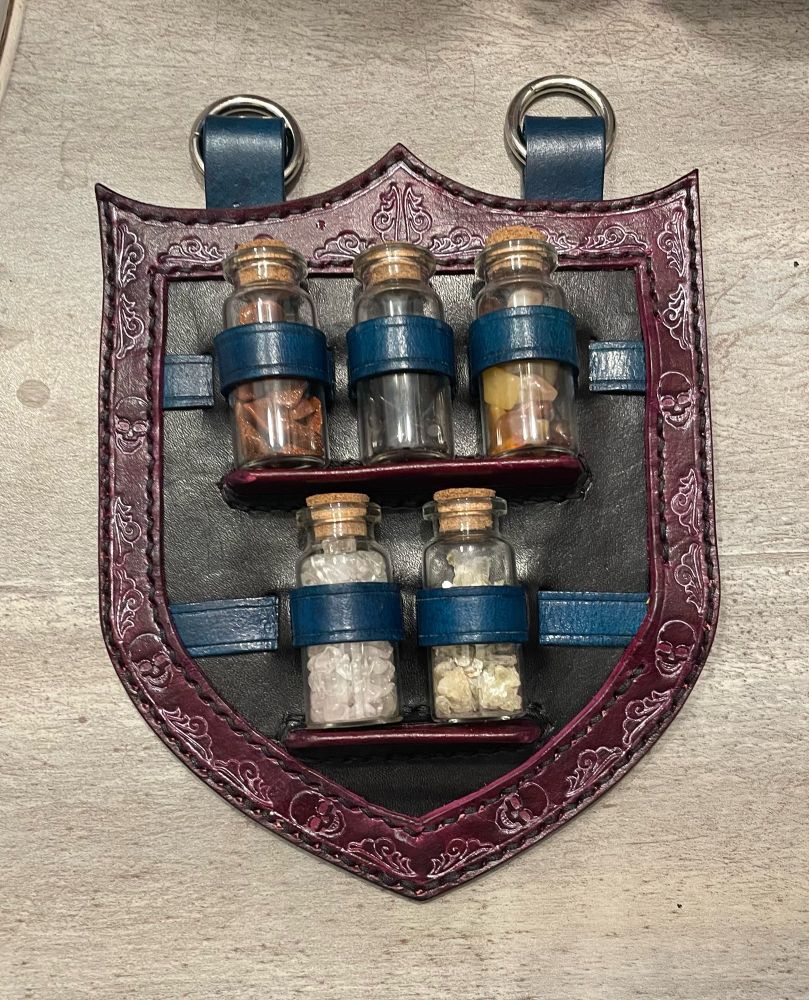A potion bottle holder that holds 5 bottles, in the shape of a shield, oxblood border with skulls and flourishes and turquoise straps on a black background. 