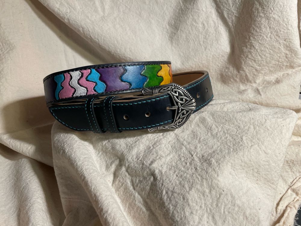 A black belt adorned with the colours of the pride and trans flags, done with squiggly lines (because it’s not about being straight, hehehe) the belt has turquoise stitching and a buckle with Celtic style pattern on it. 