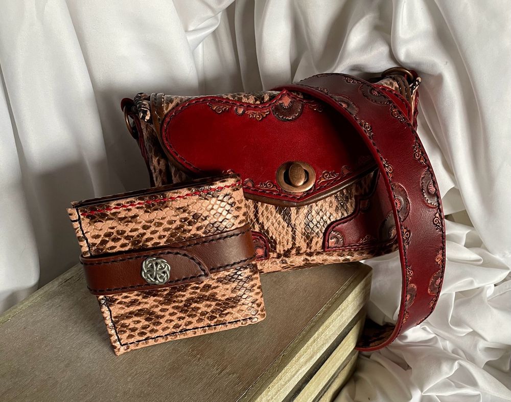 A light coral and bronze snake skin embossed wallet with a brown butter leather strap and a silver Celtic knot snap closure rests against a coordinating purse that has red leather accents that have been tooled and stamped, painted with copper and coral.
