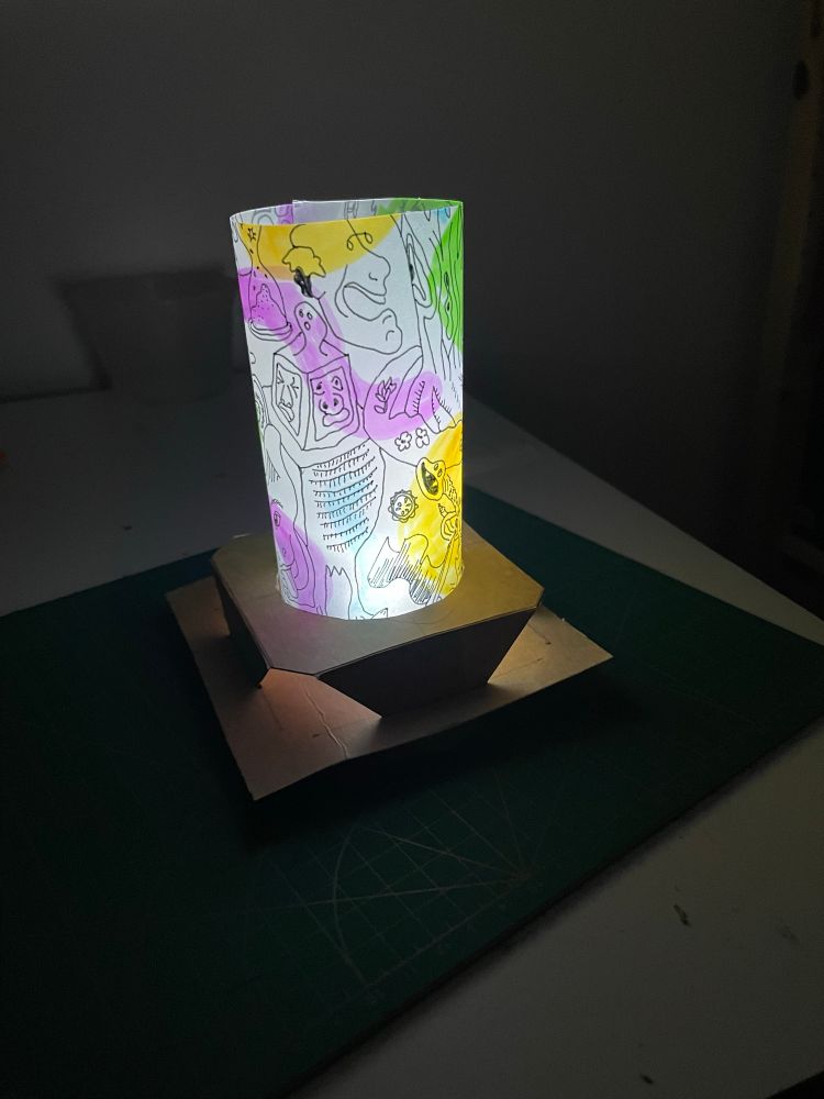 A lamp made of cardboard and translucent paper with drawings on it, lighting up a dark room