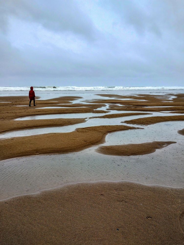 View out to sea on a rainy day, with water pooled on the sand, and a person walking on the left of the photo. 