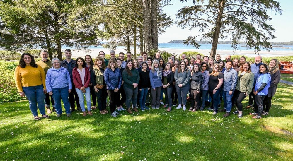 A large group of people from nature united standing on a grassy area with a beach and ocean behind them.