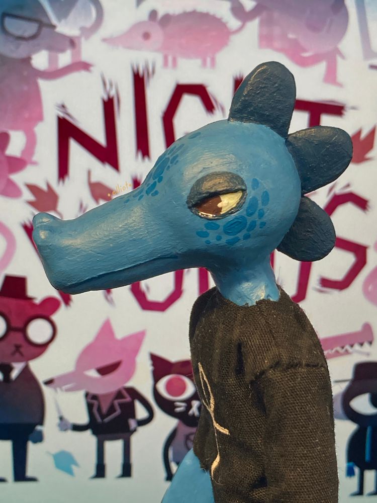 Picture of a clay doll, Bea from the game Night in the Woods, zooming in on her face. She’s a blue crocodile with a dark crest, wearing a black dress