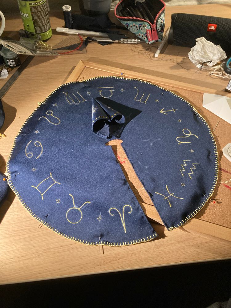 Picture of a blue cloak with golden zodiac signs painting in golden paint. It’s on its way to be finished, the hood is waiting to be sewn