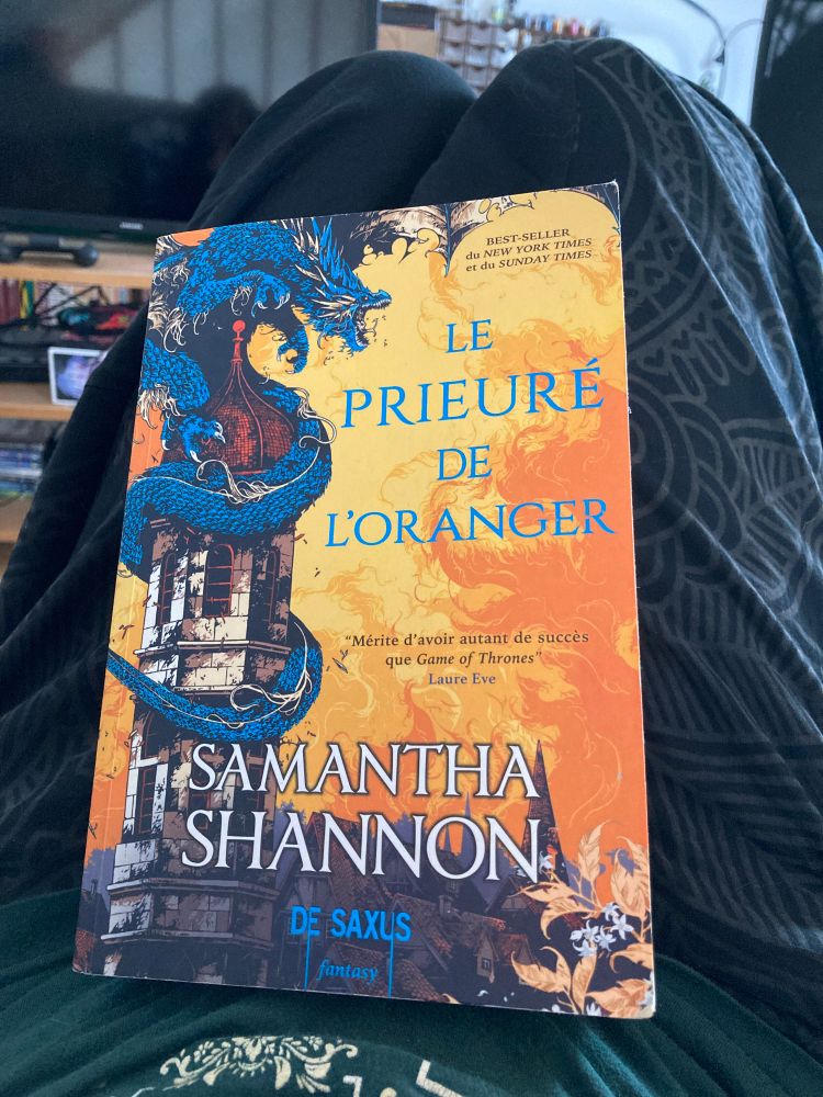 Picture of a Fantasy book « Le prieuré de l’oranger » by Samantha Shannon. The cover is burning orange with a blue dragon around a tower, with some stupid review saying it’s as good as Game of Thrones when those shouldn’t even be compared, because they’re not the same ambiance at all and there’s so much more variety to Fantasy than just being similar to GoT