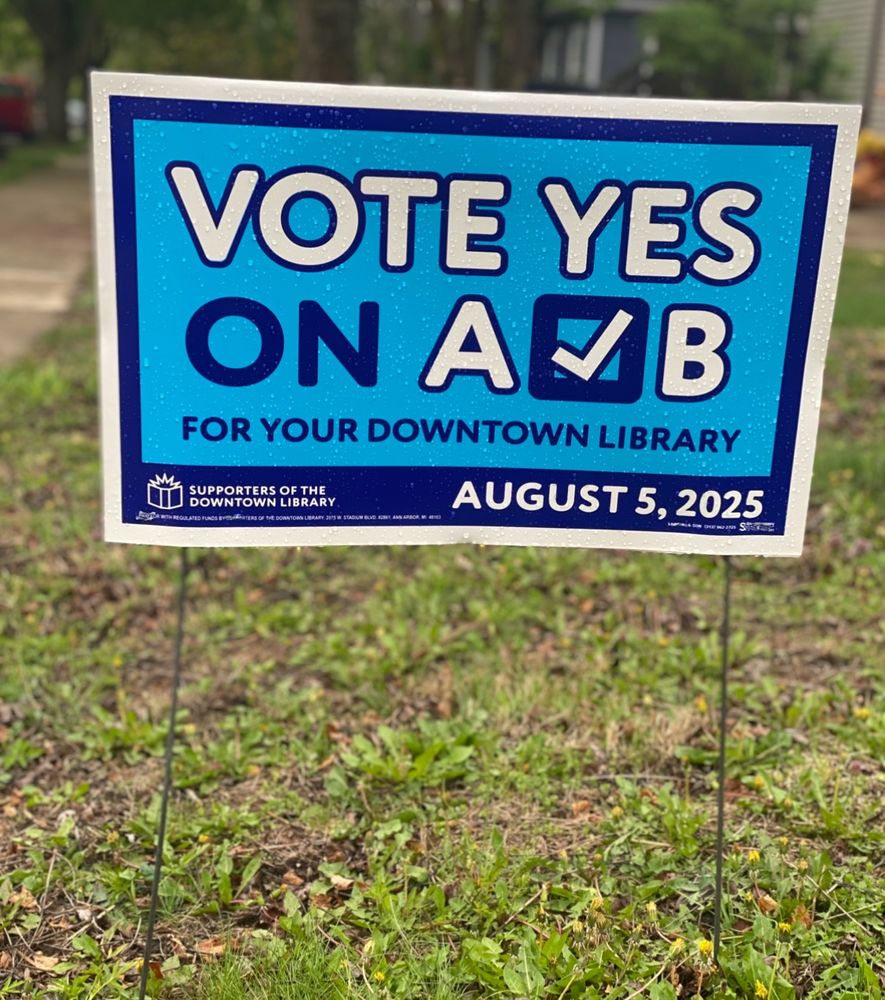 A cheerful blue yard sign that says “VOTE YES ON A & B for your downtown library. August 5 2025.” except the ampersand is a check box 