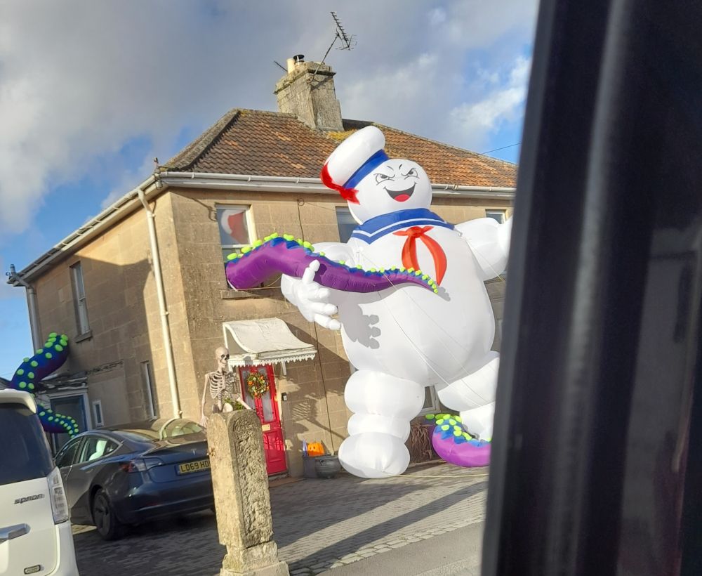 2 storey house with huge evil inflatable Mr Staypuff outside (hat above the gutter height), and purple inflatable tentacles with lime green suckers extruding out of windows.
Out of sight in front of Mr Staypuff is a small Ghostbuster in beige jumpsuit. 