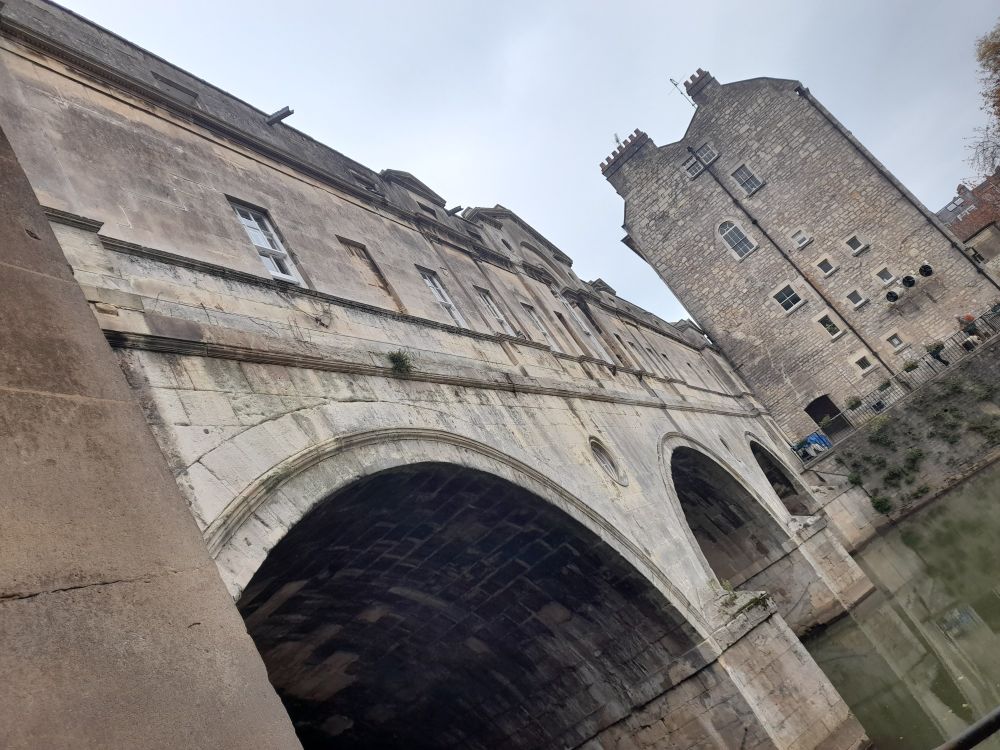 A view of Pulteney Bridge, Bath from an angle below (taken from the colonnades next to the river). 3 arches (with side angle view of the shops & their windows above) are visible with some reflection in the water, decreasing in size leading to the side of a 4-5 storey building on the opposite bank which is taller than the bridge.