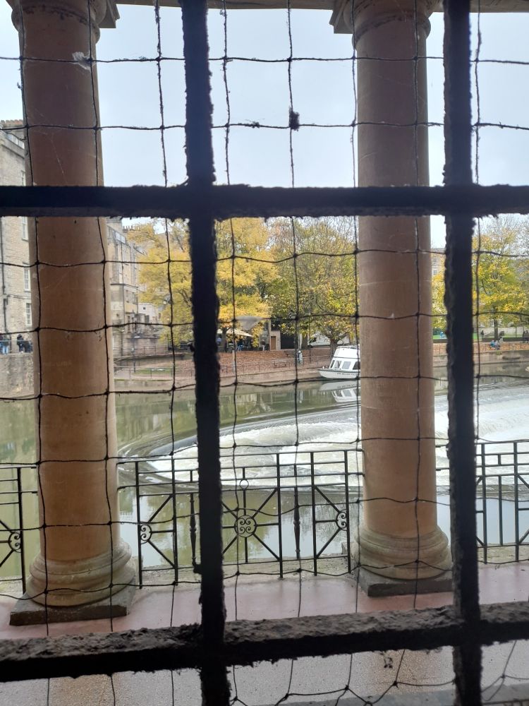 View of Pulteney Weir, looking through window bars and metal mesh in the under-road vaults, that look out between 2 of the Colonnades' columns alongside River Avon, Bath. 