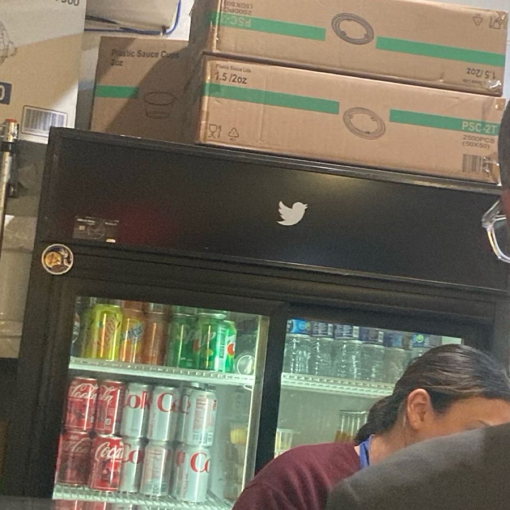 A glass door refrigerator full of soda cans behind the counter at a taquera with a white Twitter bird silhouette on it
