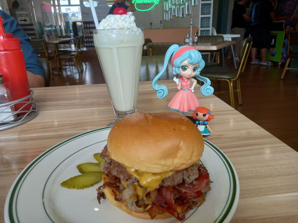A 1950s-inspired Miku Hatsune figurine and 1950s diner waitress Lalaloopsy doll put next to a milkshake and bacon cheeseburger.