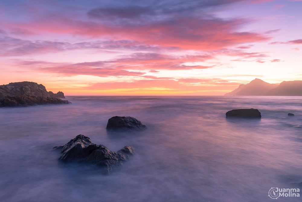 Fotografía al atardecer en la bahía de Portman. Larga exposición con filtro ND y degradado suave para lograr una velocidad de obturación lenta y unas luces compensadas en toda la escena. En primer plano el temporal 'suavizado' con el efecto seda debido a la larga exposición, con unas piedras que conducen hacia el horizonte. Las nubes tienen un color rosado y anaranjado debido a la puesta de sol.