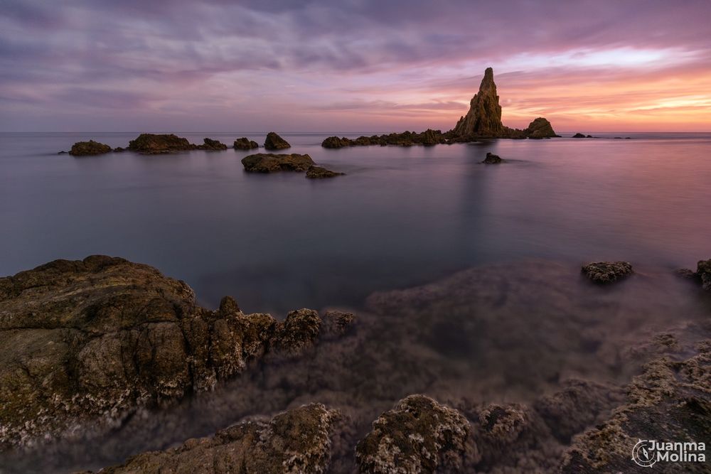 Fotografía de larga exposición al atardecer, con el agua efecto seda que hace ver qué está en absoluta calma. Con unas rocas en primer plano que llevan la mirada hasta el hito de piedra situado en un plano más alejado. Todo ello bañado con la luz anaranjada del atardecer
