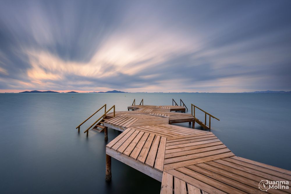 Fotografía de larga exposición al amanecer. En la mitad inferior tenemos un muelle con formas hexagonales con algunas escaleras que entrar en el agua con efecto suave gracias a la larga exposición. El cielo está marcado por nubes a la fuga en movimiento en dirección al Sol que comienza a aparecer en el horizonte.