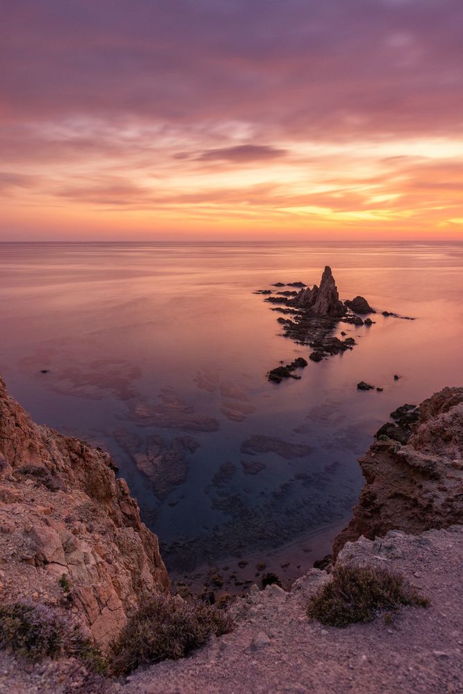 Fotografía de larga exposición al atardecer, con el agua efecto seda que hace ver qué está en absoluta calma. Con unas rocas en primer plano que llevan la mirada hasta el hito de piedra situado en un plano más alejado. Todo ello bañado con la luz anaranjada del atardecer