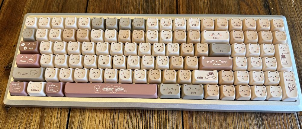 Photo of a full aluminum keyboard with keycaps that are shaped like they have tiny cat ears. The keys are mostly off-white with some beige, grey, a brownish-red and some brown keys. Most keys have the face and whiskers of a cat on them in addition to the key legend. Others have symbols for coffee cups and beans. The spacebar has a logo that reads “Coffee Kitty.”