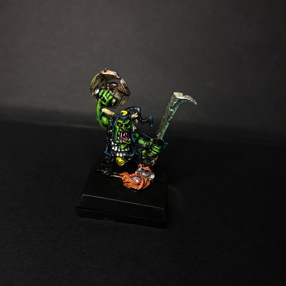 A happy Goblin stands on the head of a less happy Dwarf. The Goblin has a shield lifted in triumph and is manically leering 